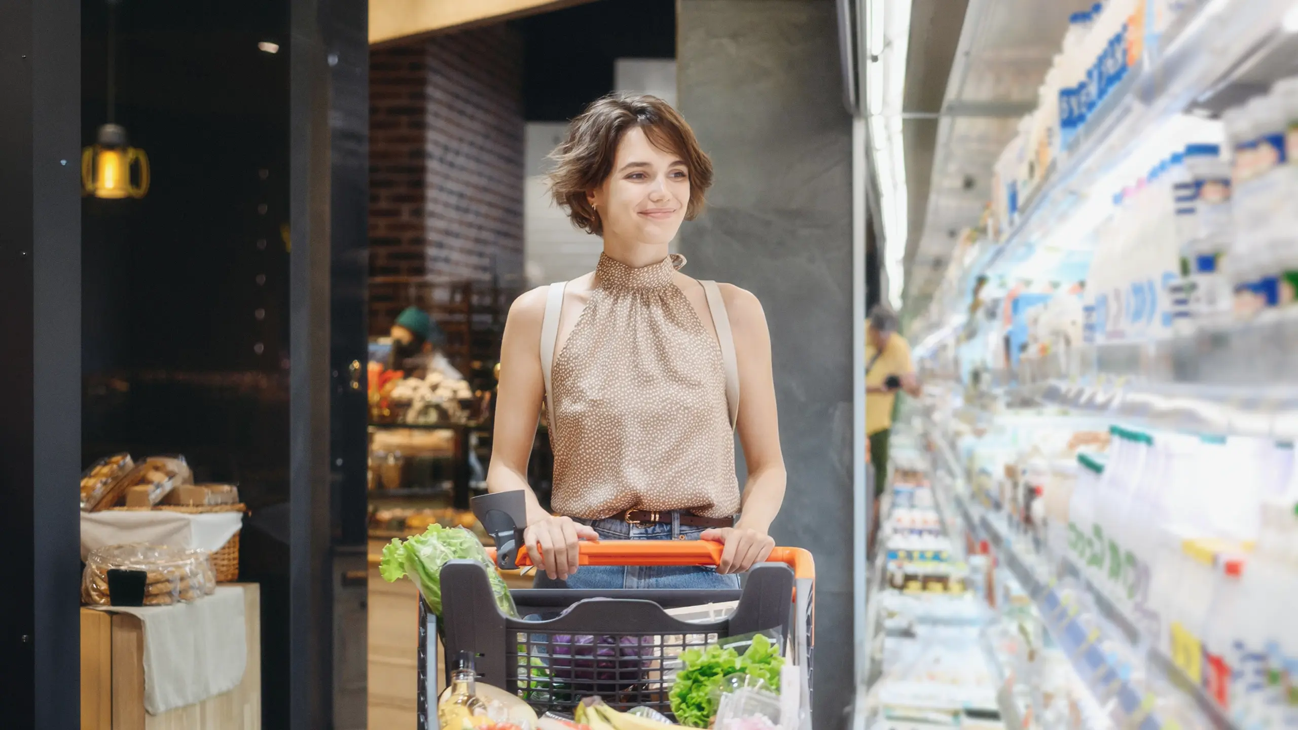 A shopper pushing a cart through a grocery store, comparing products on a bright, blurred aisle.